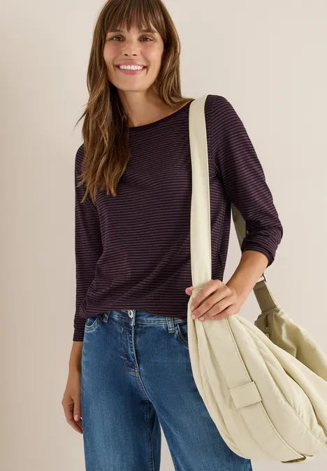 Frau mit dunklem Shirt, Jeans und heller Umhängetasche lächelt.